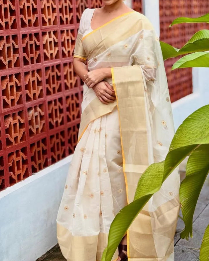 Woman in a beige saree standing in front of a decorative brick wall.
