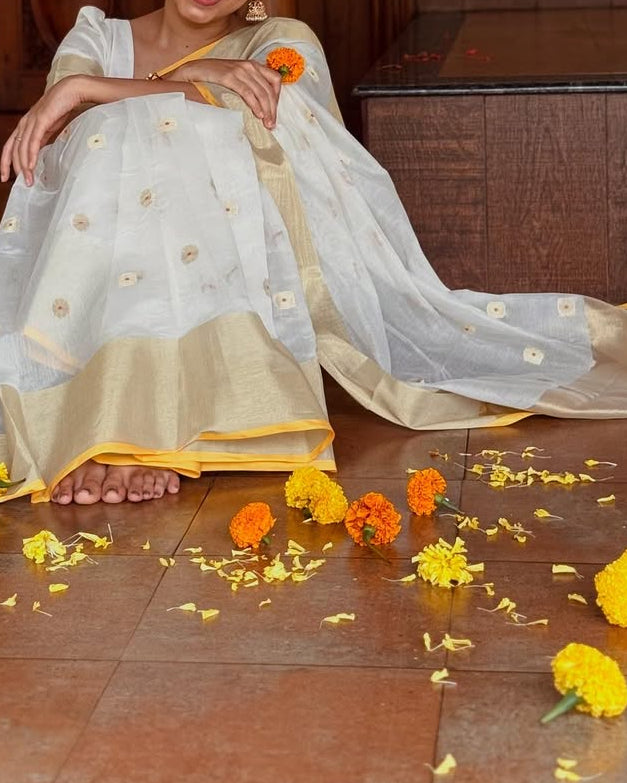 Woman in a white and gold traditional outfit sitting on a wooden floor with flowers around her.