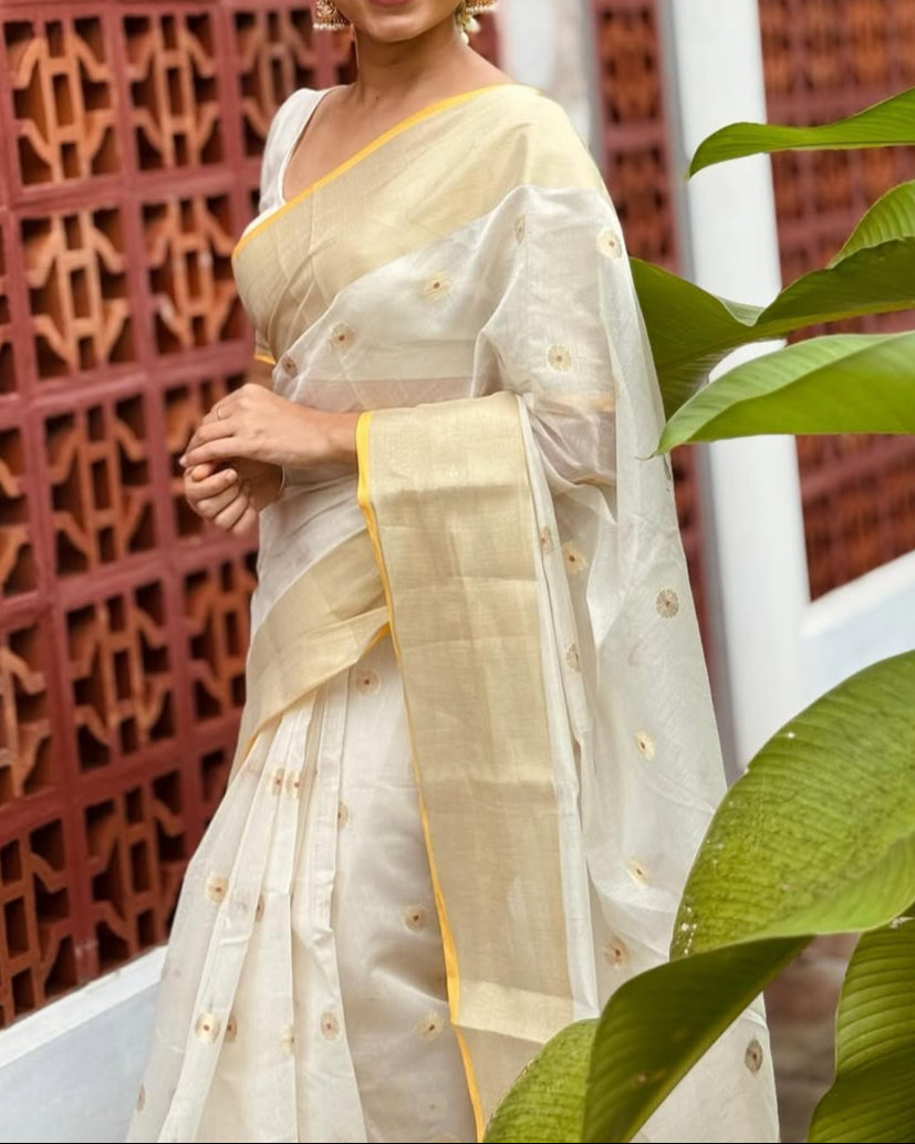 Woman in a white saree with a yellow border standing against a textured red wall.