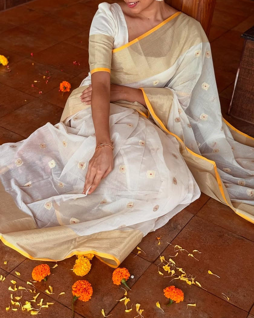Woman in a white saree with yellow trim sitting on a wooden floor surrounded by flowers.
