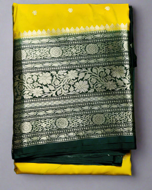 A folded Banarasi silk saree with yellow and green colors and intricate silver Zari weave designs on the border and pallu, displayed on a green grass-like surface.