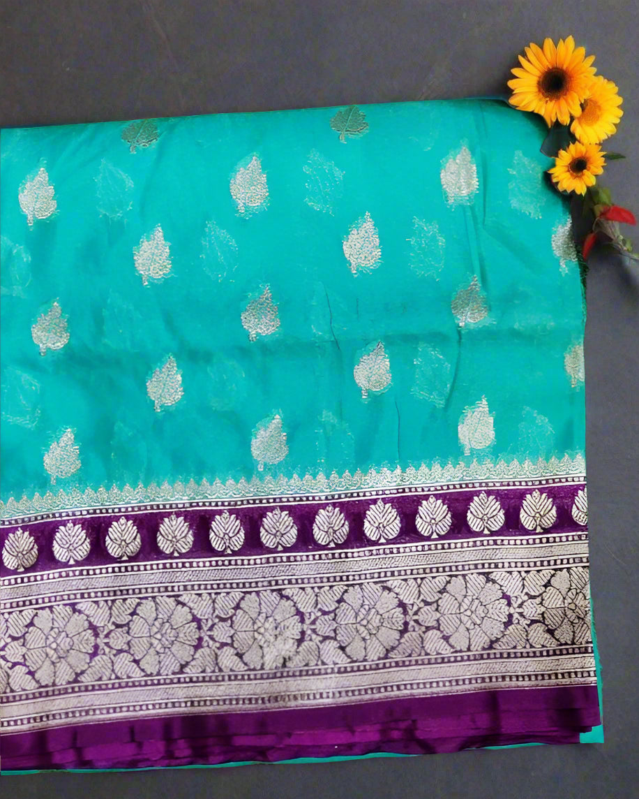 A folded Handwoven Banarasi Katan Georgette Saree in blue and purple, adorned with intricate buti designs and Zari weave on the pallu and blouse, placed on a grass-like surface with flowers in the background.