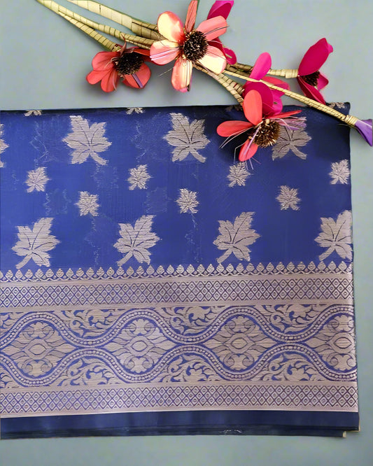 A blue saree with floral and paisley patterns displayed on a grassy surface, alongside artificial flowers.