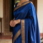 Woman in a blue saree with gold border standing in an indoor setting.