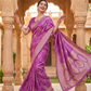 Woman in a purple saree standing in front of an architectural background