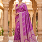 Woman in a purple saree with intricate patterns standing in an architectural setting.