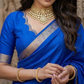 Woman in a blue saree with gold jewelry standing in front of stone architecture.