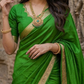 Woman in a green saree with gold border standing in front of stone architectural background