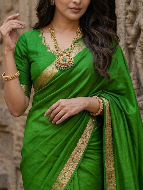 Woman in a green saree with gold border standing in front of stone architectural background
