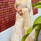 Woman in a beige saree standing in front of a decorative brick wall.
