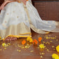 Woman in a white and gold traditional outfit sitting on a wooden floor with flowers around her.