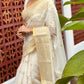 Woman in a white saree with a yellow border standing against a textured red wall.