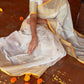 Woman in a white saree with yellow trim sitting on a wooden floor surrounded by flowers.