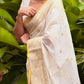 Woman in a white saree with yellow accents standing among green leaves