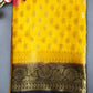 A yellow Khaddi Georgette saree with gold zari weaving displayed on a green surface, with a decorative border at the pallu.