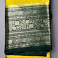 A folded Banarasi silk saree with yellow and green colors and intricate silver Zari weave designs on the border and pallu, displayed on a green grass-like surface.