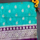 A folded Handwoven Banarasi Katan Georgette Saree in blue and purple, adorned with intricate buti designs and Zari weave on the pallu and blouse, placed on a grass-like surface with flowers in the background.