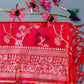 A red heavy silk saree with intricate Zari and Minakari weaving, displayed on a grassy surface with a few flowers at the top right corner.