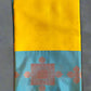 A folded Yellow Banaras Kora Muslin Saree with intricate designs and Zari weave on the pallu and blouse, displayed on a green background.